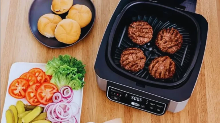 Four grilled burger patties inside the Ninja AG301 with buns, lettuce, tomatoes, pickles, and onions on a wooden table
